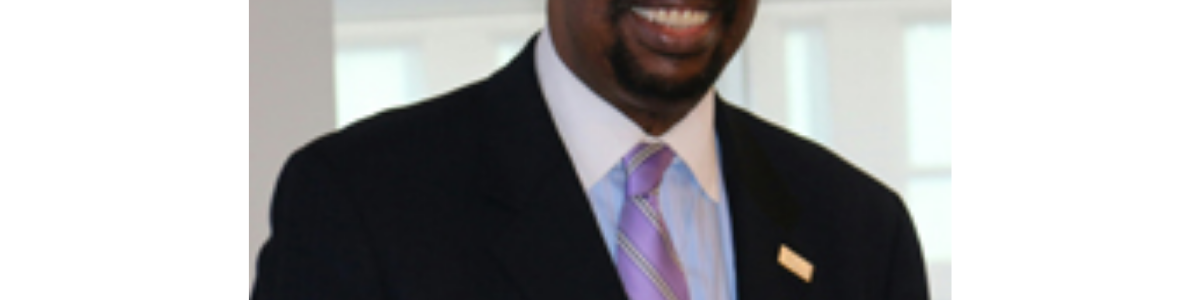 Black man smiling in black suit jacket, blue shirt with white collar and lavender tie