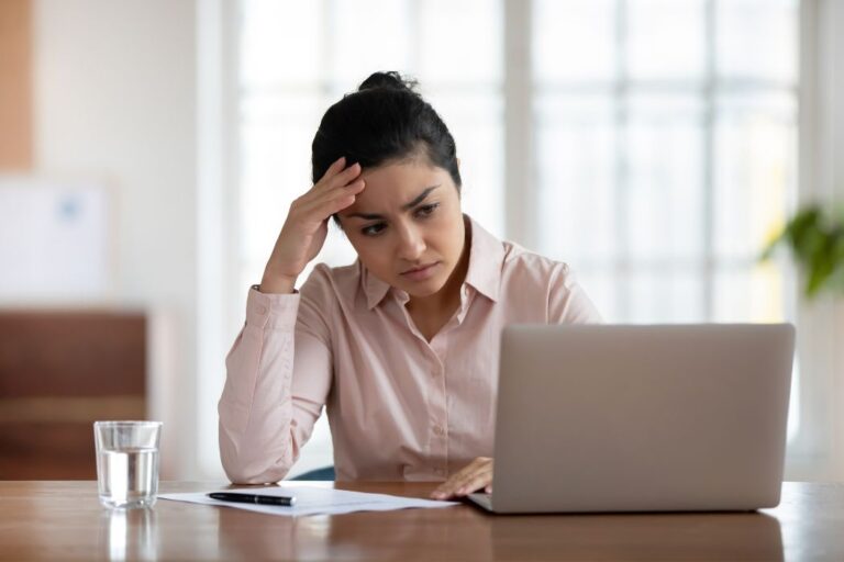 Exasperated woman sitting at desk with laptop holding her head in her hand.