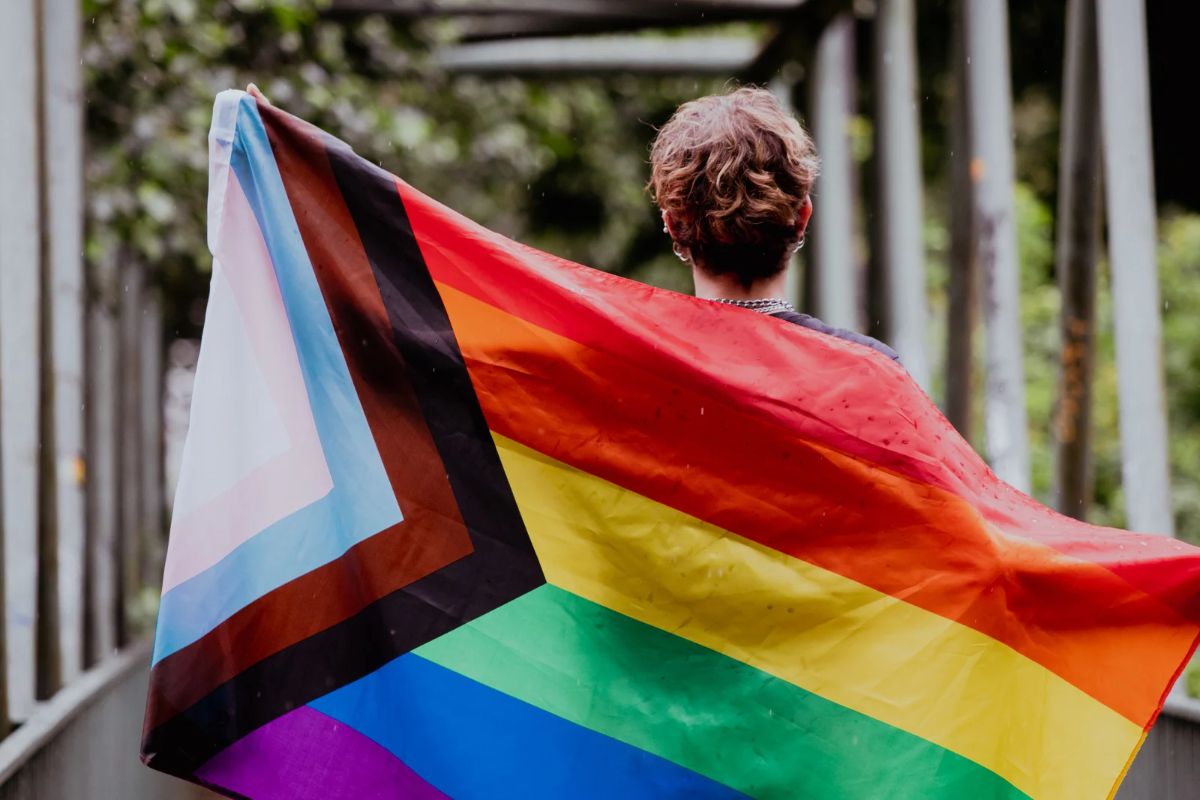 Man standing with his back to the camera with a Pride flag draped across his back.