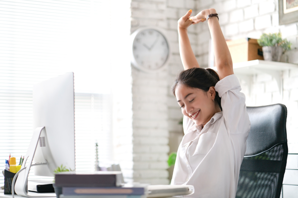 Young Asian woman sitting at desk with her arms raised in the air stretching.
