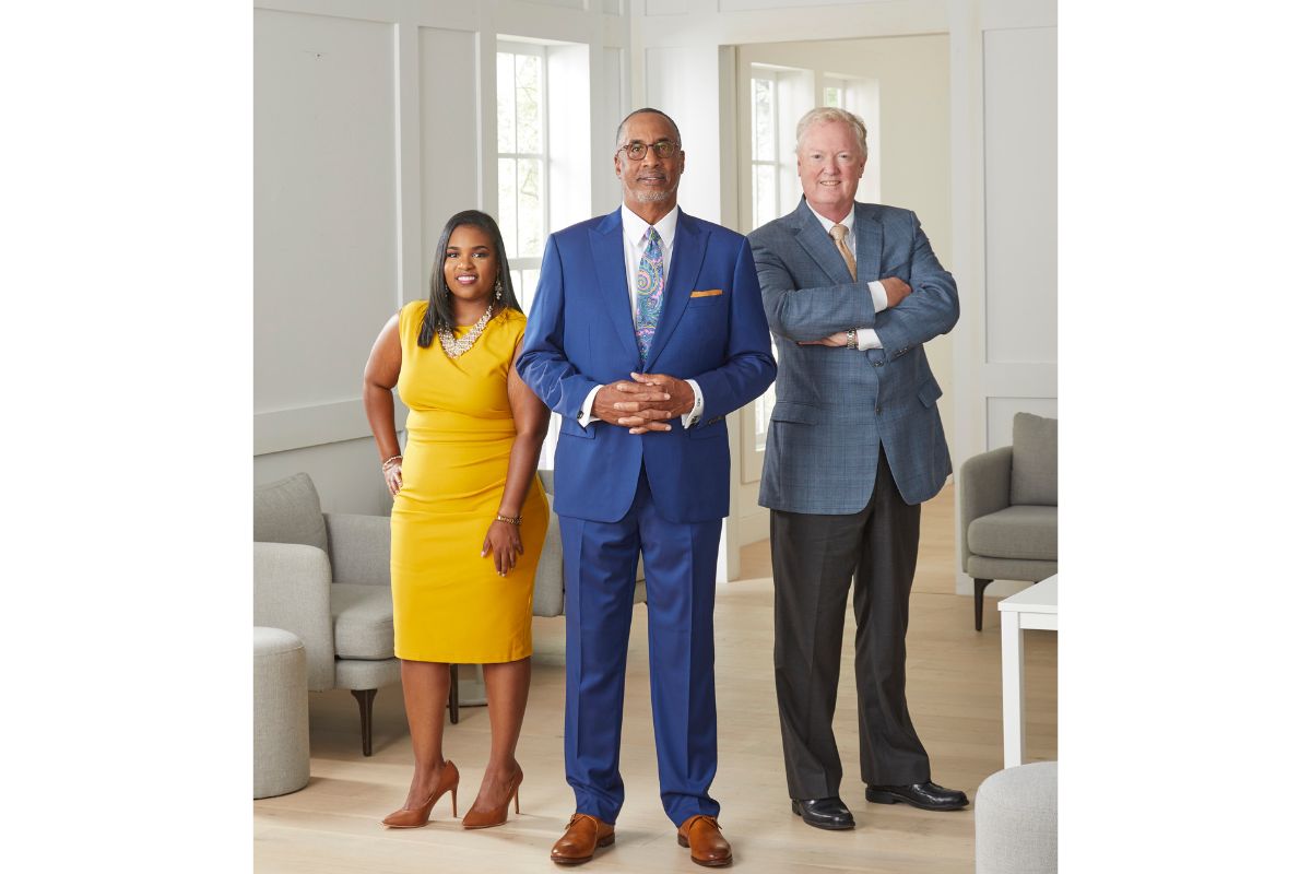Black in blue suit standing in a room with a white man on his left and a Black woman in a yellow dress on his right.