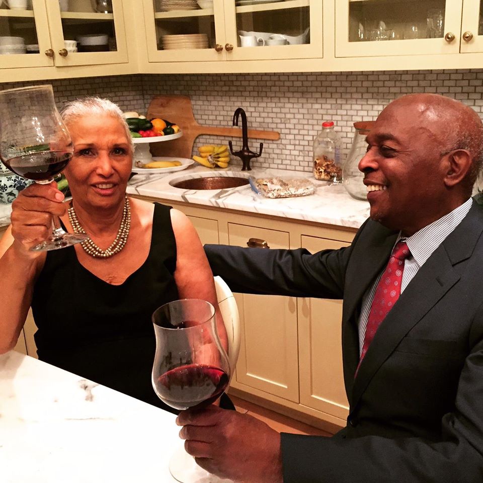 An older Black couple sitting at a kitchen table holding wine glasses.
