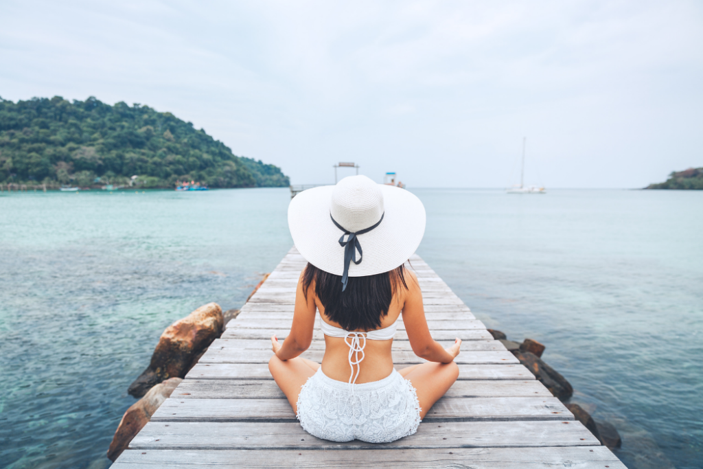 Woman sitting on a pier in a white beach hat and white bikini with her back to the camera.