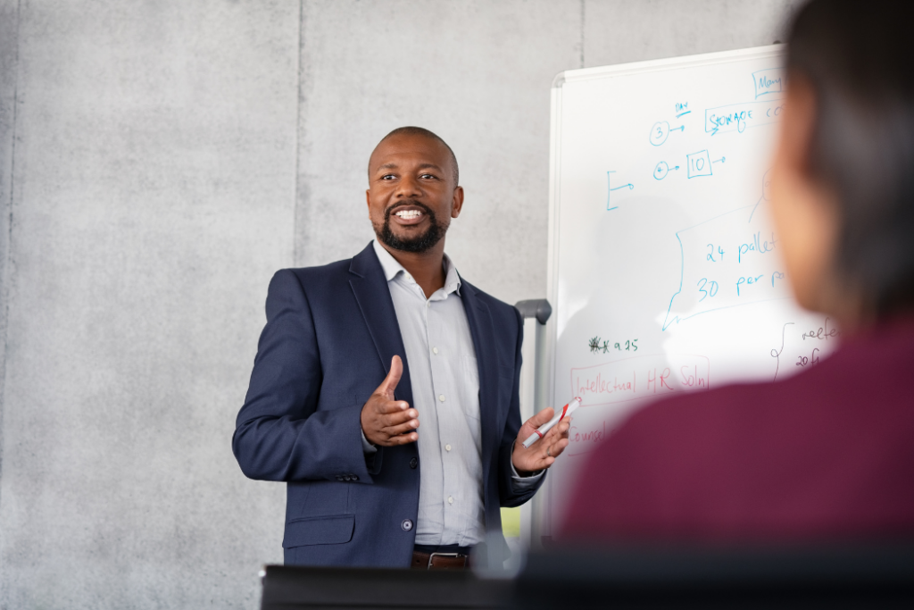 Black man in white shirt and blue jacket stands in front of a whiteboard delivering an address.