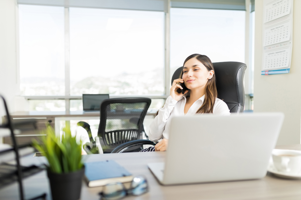 Dark-haired woman sits at desk talking on the phone.