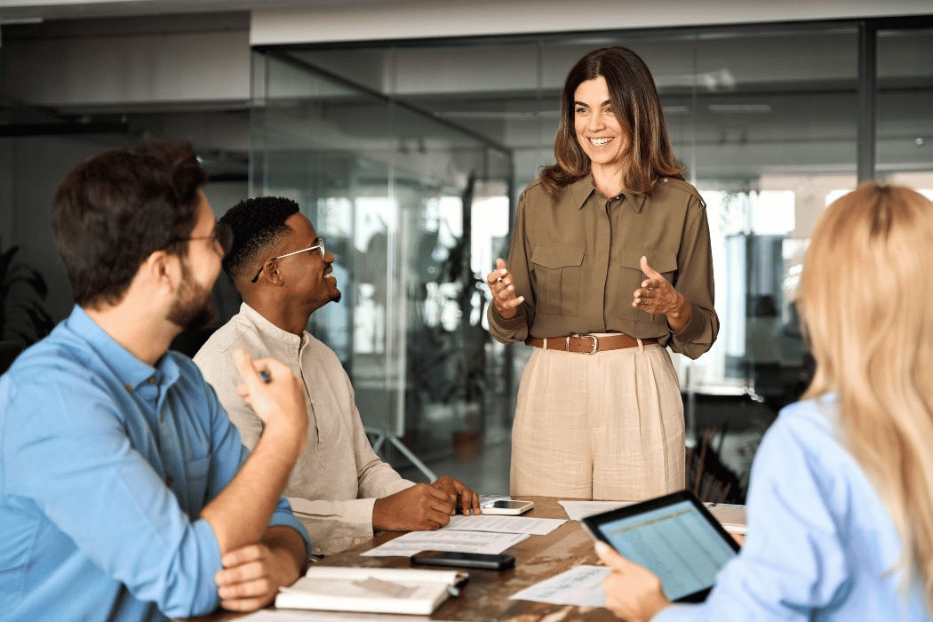 Woman stands addressing three people seated at a conference table.