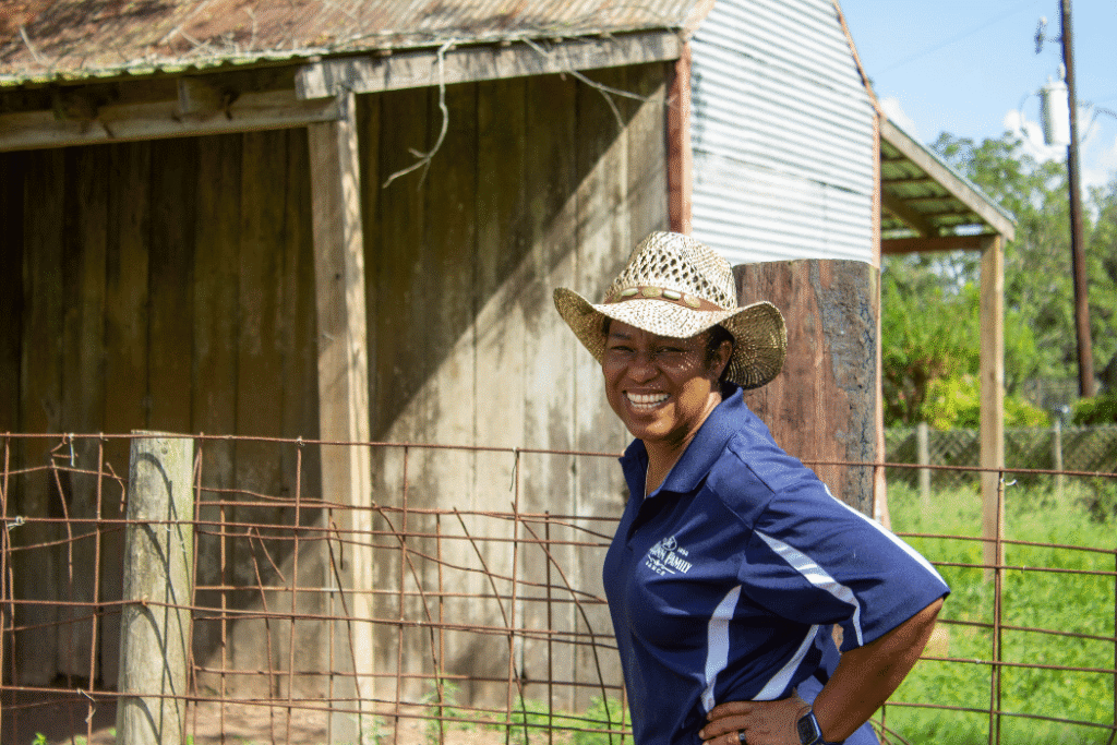 A Black woman in a cowboy stands in front of a barn smiling with her hand on her hip