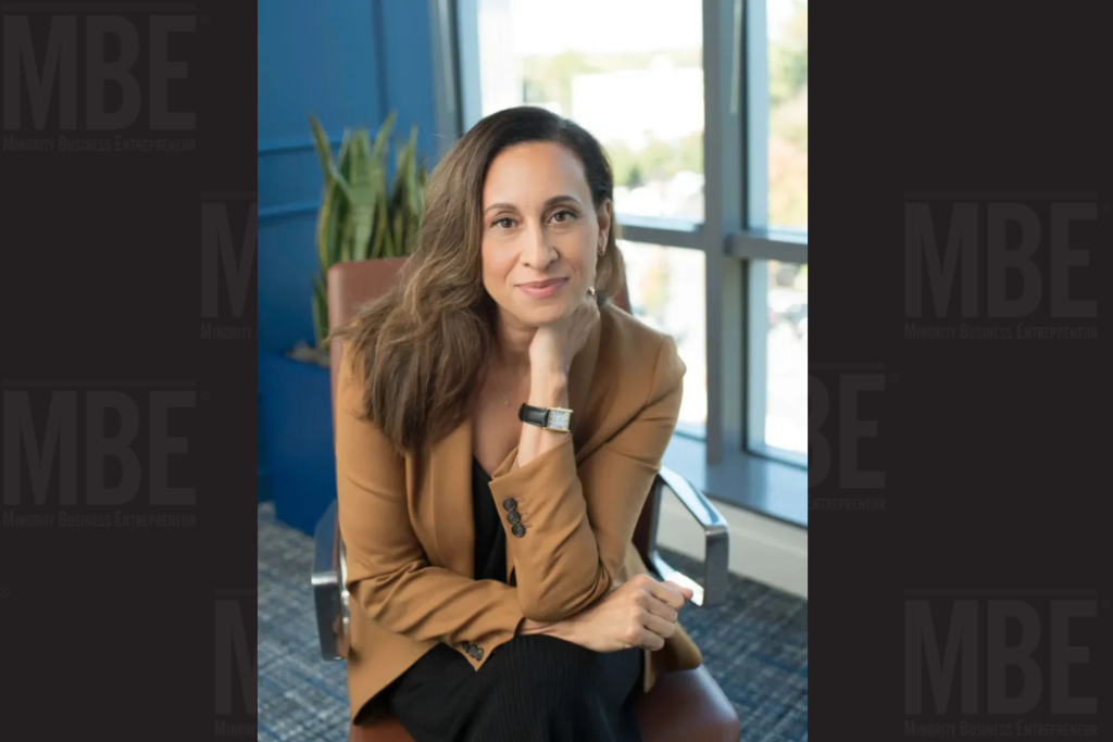 Headshot of Britt Hogue in a brown jacket with one hand across her knee and the other under her chin