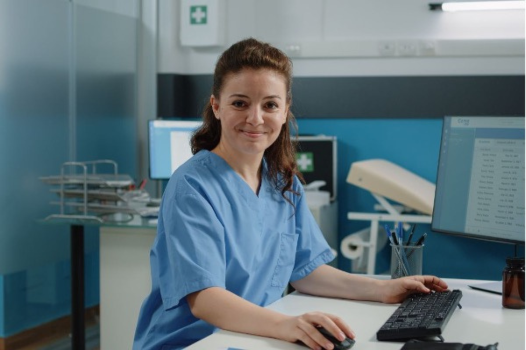 A nurse smiling at the camera and sitting at a desk using a computer