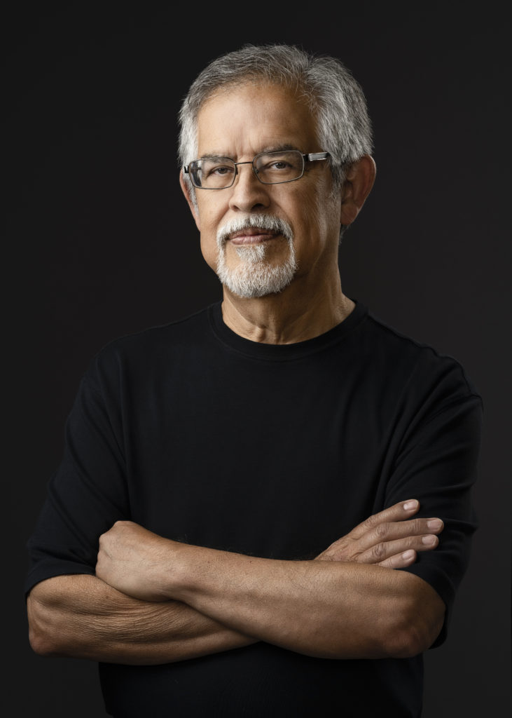 Photo of latino man with gray hair standing with his arms folded against a black background.