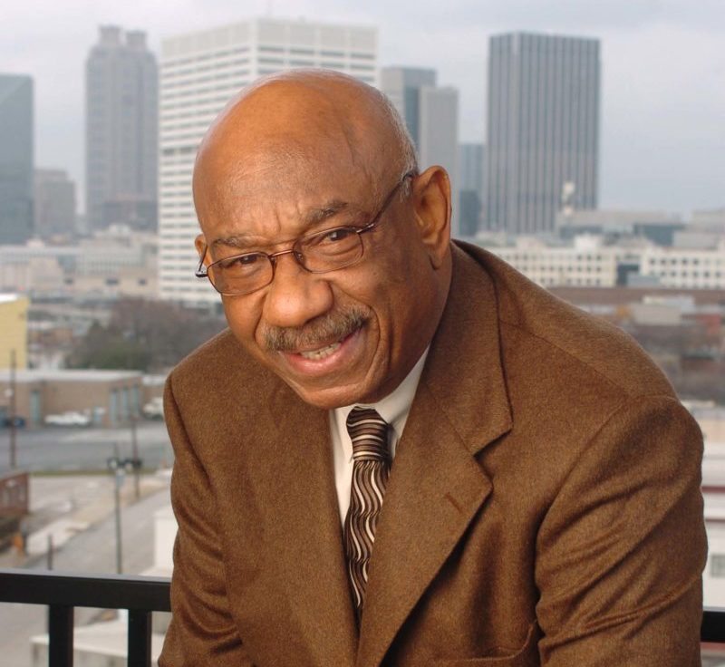 Photo of entrepreneur Herman J. Russell leaning on his knee on a balcony with the Atlanta skyline behind him.