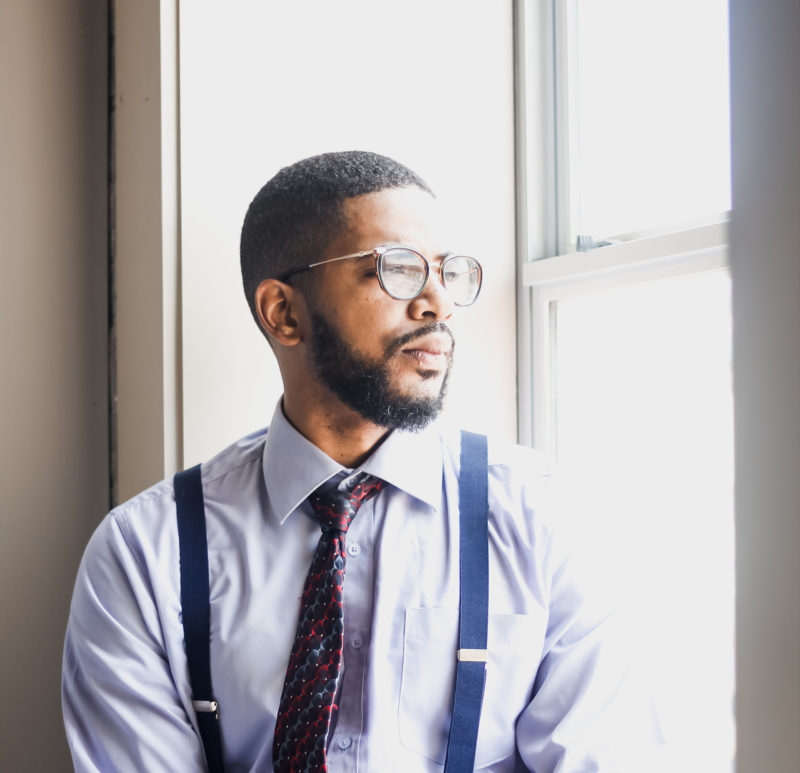 Professional black man looking lonely while sitting on window sill looking out.