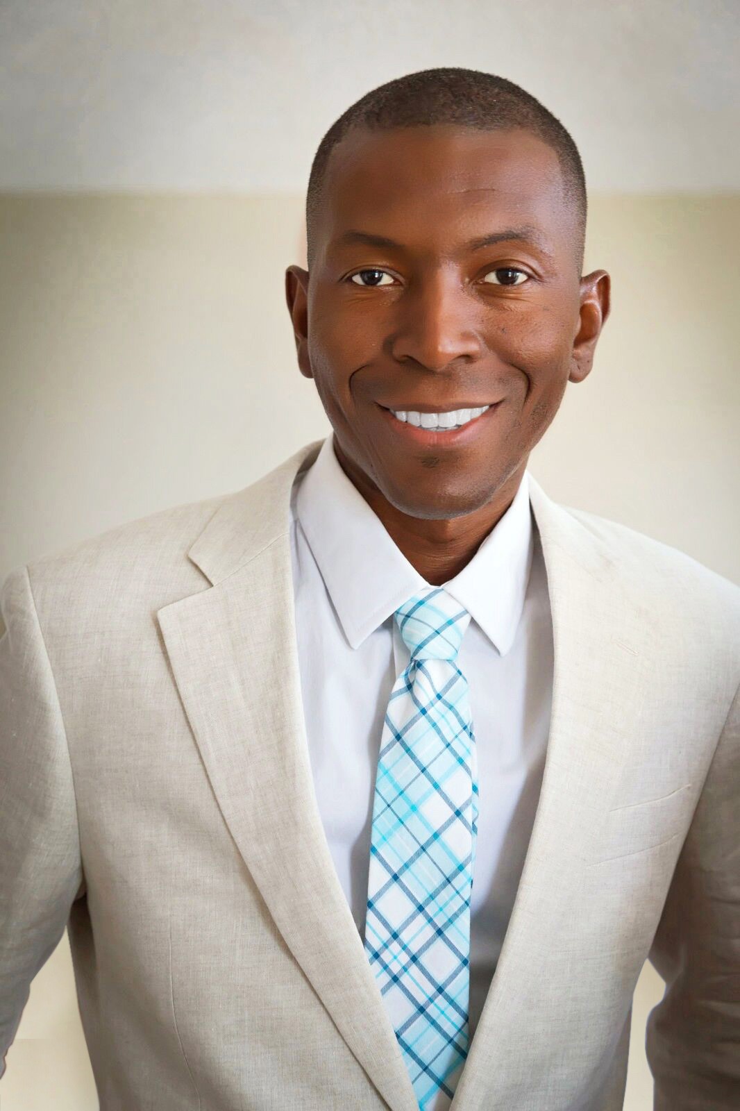 A black man in a cream suit with a royal blue plaid tie smiling and looking at the camera.